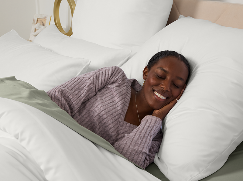 Frau schläft friedlich in einem Bett mit weißen und salbeigrünen Bettlaken, trägt einen lavendelfarbenen Pullover und lächelt entspannt.