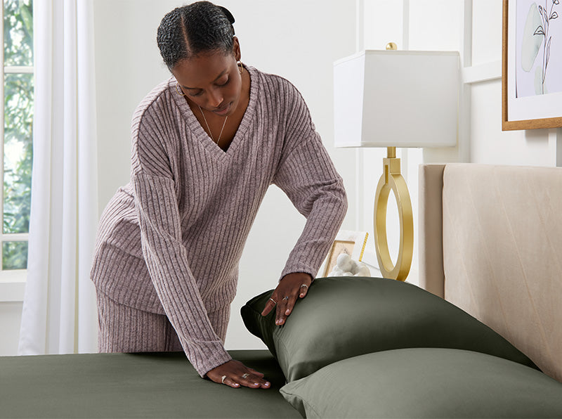 Frau in lavendelfarbener Loungewear glättet salbeigrüne Bettlaken auf einem ordentlich gemachten Bett in einem hellen, modernen Schlafzimmer.