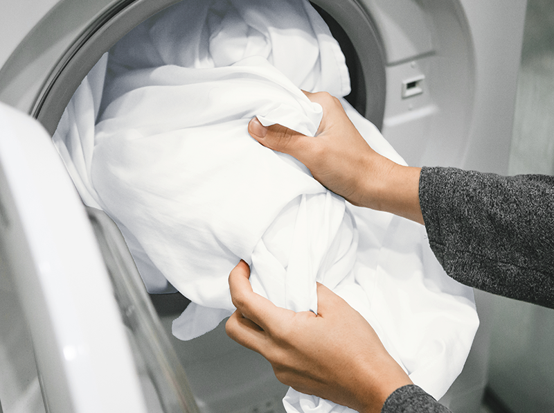 Hände legen weiche weiße Bettlaken in die Waschmaschine, ideal für einfache Pflege und Maschinenwäsche.